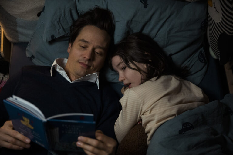 Standbild aus dem Film "Eine Milion Minuten": Wolf (Tom Schilling) liest seiner Tochter Nina (Pola Friedrichs) im Bett ein Buch vor.