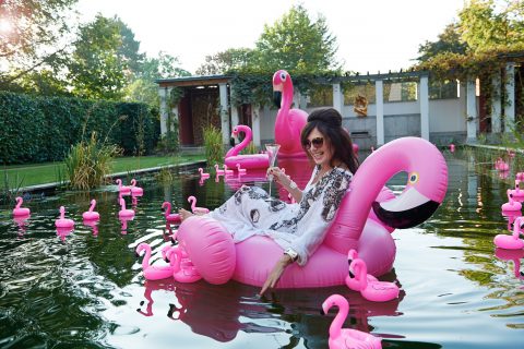 Eine Frau sitzt lachend auf einem großen, pinkfarbenen Flamingo-Schwimmreifen in einem Teich, umgeben von vielen kleineren pinken Flamingo-Schwimmfiguren. Sie trägt eine helle, gemusterte Tunika, Sonnenbrille und hält ein Sektglas in der Hand, während sie mit der anderen Hand das Wasser berührt. Im Hintergrund ist ein Garten mit Büschen, Bäumen und einer Pergola zu sehen.