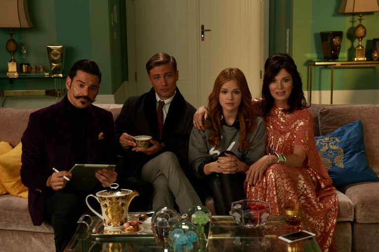 Standbild aus dem Film "High Society": Von links nach rechts sitzen Olivjeh (Rick Kavanian), der PR - Berater der Familie Schlacht, , Anabel (Emilia Schüle) und Trixi von Schlacht (Iris Berben) auf dem Sofa.
