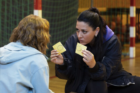 Standbild aus dem Film "Einfach mal was Schönes": Karla (Karoline Herfurth) und Senay (Jasmin Shakeri) sitzen auf einem Turnsaalboden. Senay zeigt Karla zwei Zettel. Sie soll sich entscheiden.