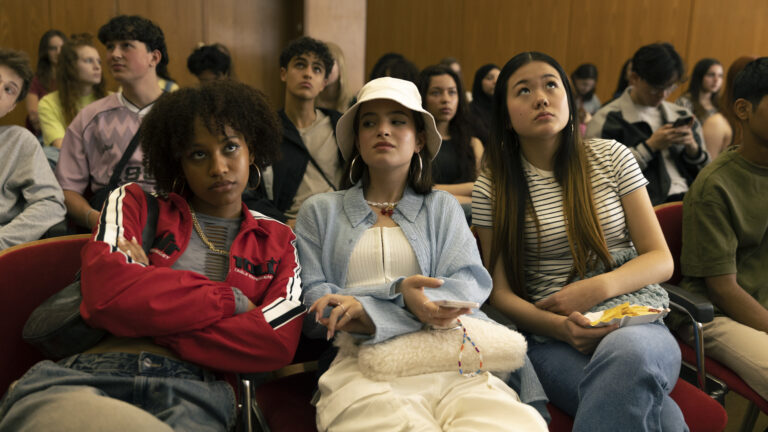 Standbild aus dem Film "Wunderschöner": Die drei Freundinnen – Alisha (Nadia Schaffer), Lily (Emilia Packard) und Phuong Anh (Sea Starzacher) – sitzen im Publikum des voll besetzten Schulsaals.